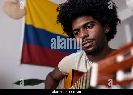 Portrait of confident serious young man with afro hair practicing guitar while sitting against Colombian flag at home Stock Photo