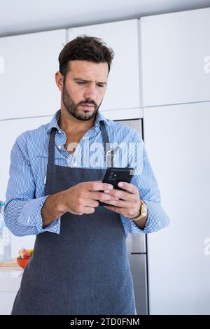 Adult bearded male chef in blue shirt and apron looking at screen of mobile phone while standing in modern kitchen and browsing for text messages Stock Photo