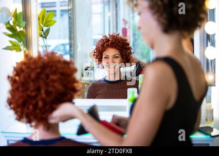 A cheerful woman with curly red hair is styling her hair using a straightener in a bright, modern hair salon, with reflections in the mirror. Stock Photo