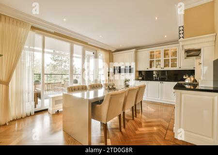 Dining table and chairs arranged in front of open plan large kitchen next to bright window in contemporary apartment Stock Photo