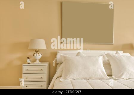Interior of bedroom with white bed and blank frame mounted on beige wall with lampshade and clock on dresser at home Stock Photo