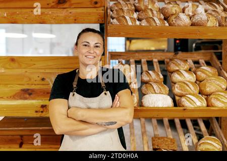 Portrait of confident positive female seller in apron with arms crossed standing by freshly baked breads arranged on wooden shelves in bakery Stock Photo