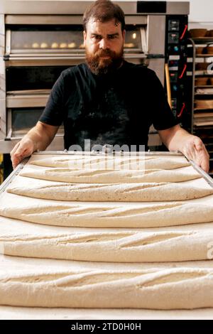 Red beard male baker in black t-shirt preparing to put tray with raw dough in oven for baking loaves of bread in kitchen at bakehouse Stock Photo