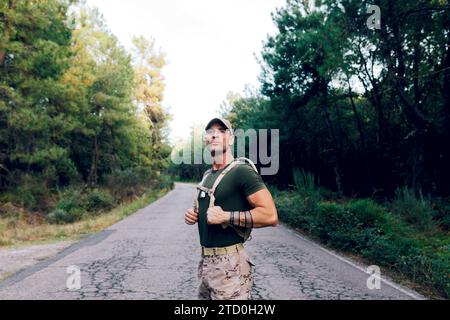 Confident mature soldier wearing camouflage pants and green t-shirt looking away while standing on road against trees in forest Stock Photo