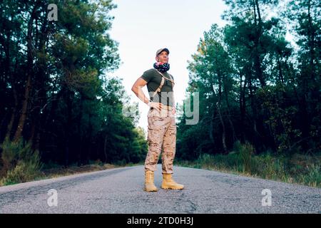 Full body of Confident middle aged military soldier wearing green t-shirt and cap with American flag wrapped around neck standing on road in forest Stock Photo