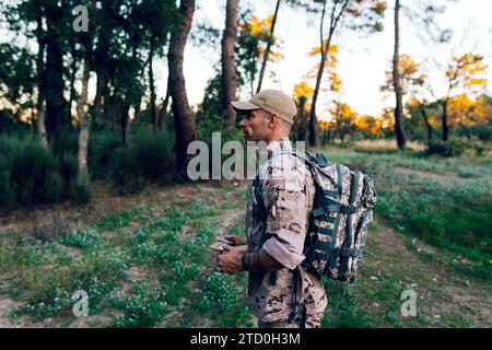 Side view of mature special force military soldier wearing camouflage uniform with cap and backpack holding box standing in forest at sunset Stock Photo
