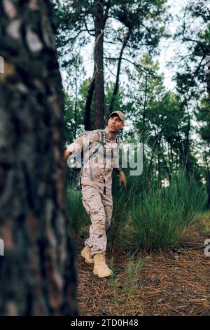 Full body of mature special force military soldier wearing camouflage uniform with cap and backpack looking away while walking in forest Stock Photo