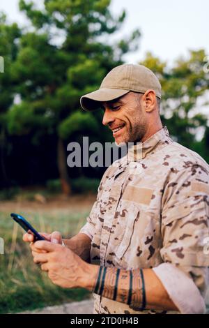 Smiling middle aged special force ranger wearing camouflage uniform and cap using mobile phone in forest against blurred background Stock Photo