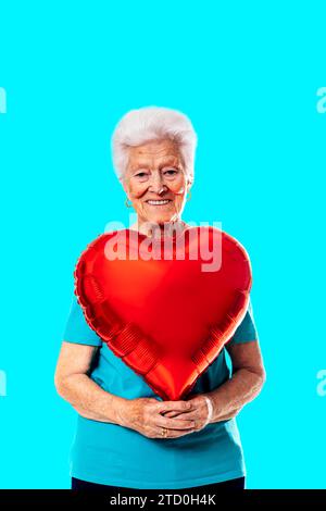 Positive elderly gray haired lady holding red heart shaped balloon and looking at camera against blue background Stock Photo