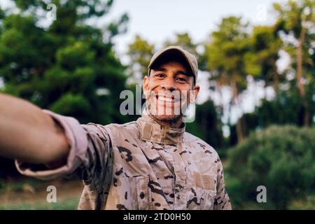 Smiling portrait of mature Caucasian special force commando wearing camouflage uniform taking selfie against trees in forest Stock Photo