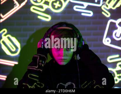 A focused boy gamer with headphones is immersed in a gaming session, surrounded by colorful neon light symbols on a brick wall. Stock Photo