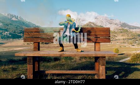 A person sits on a big wooden bench surrounded by mountains, releasing a cloud of vibrant colored smoke. Stock Photo