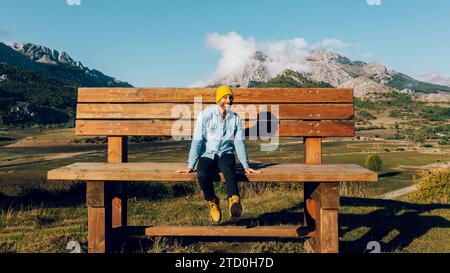A happy man in a yellow beanie and blue shirt enjoys the beauty of a mountainous landscape while sitting on a big wooden bench. Stock Photo
