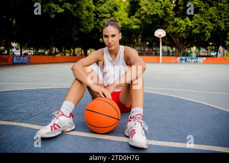 Full body of sporty young female in activewear sitting with ball and looking at camera on basketball court with blurred trees in background Stock Photo