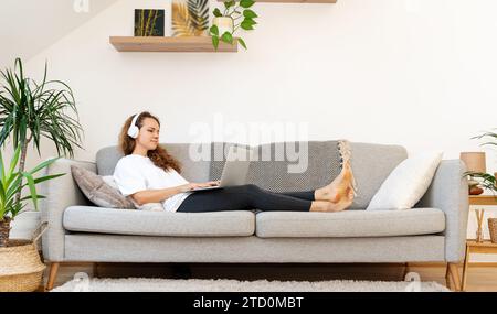 Woman with wireless headphones lying down on couch and using her laptop. Stock Photo