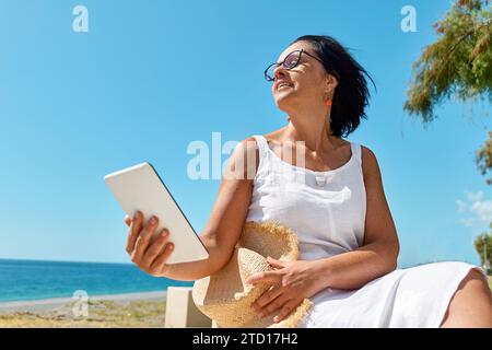 Mature woman wearing using digital tablet at the beach. Middle aged female using digital device for shopping online, banking, social media or reading Stock Photo