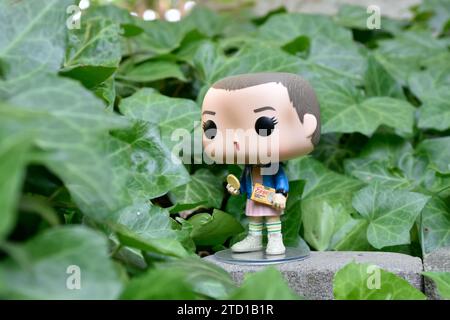 Funko Pop action figure of Eleven with Eggo waffles from Netflix TV series Stranger Things. Green ivy plant leaves, abandoned garden, mysterious mood. Stock Photo