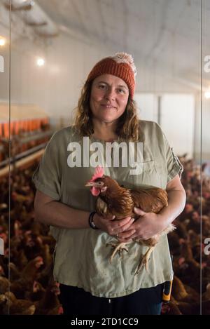 A female farmer stands holding a chicken surrounded by a flock of chickens and hens. Stock Photo
