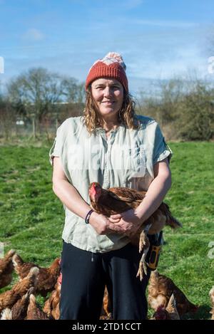 A female farmer stands holding a chicken surrounded by a flock of chickens and hens. Stock Photo