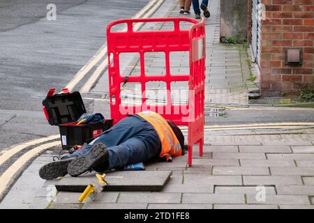 A BT Openreach engineer lying out on a pavement working on underground telephone fibre cables accessed by lifting an inspection cover on a street Stock Photo