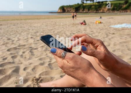A woman looks at her mobile phone while sitting on a beach in Brittany in the North of France Stock Photo