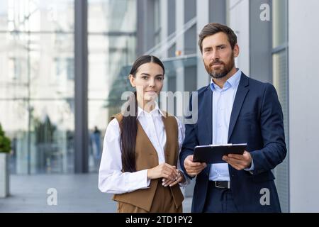 Portrait of a team and partners of a young business man and woman standing outside the office center with documents in their hands, smiling and confidently looking at the camera. Stock Photo