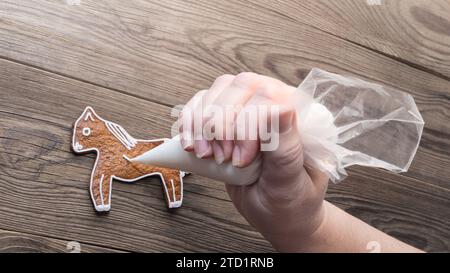 Cute gingerbread horse or human hand with piping bag on brown wooden background. Closeup of decorating sweet baked cookie in pony shape by white icing. Stock Photo