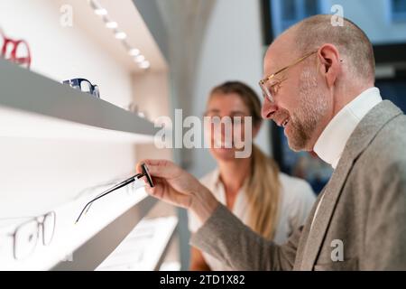 Optician and customer discussing new eyeglasses. Shelves with various frames in an optical shop. Stock Photo