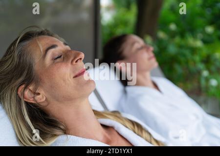 Two women relaxing on outdoor loungers, one in focus with eyes closed. Spa wellness hotel concept image Stock Photo