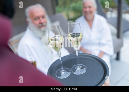 Waiter offering champagne to a smiling senior couple in white bathrobes at a hotel Stock Photo