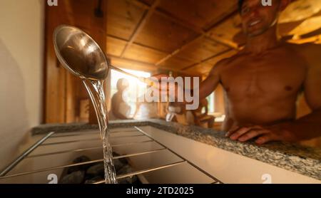 Man pouring water from a sauna ladle onto hot rocks on a sauna stove. People in the background. Finish sauna spa wellness hotel concept image. Stock Photo