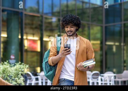 Indian young male student walking on street near campus and university with backpack. Holds books and uses a mobile phone. Stock Photo