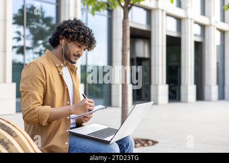 Indian young male student sitting outside on a bench and studying online, watching and listening in headphones to online lectures, writing down in a n Stock Photo