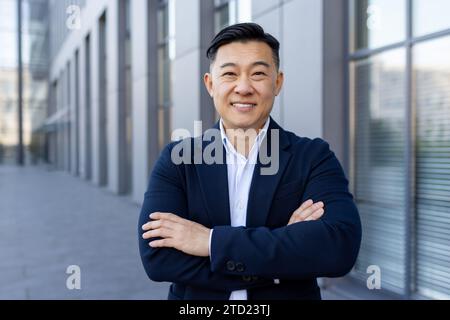 Close-up portrait of a smiling young businessman standing in a suit outside an office center, crossing his arms over his chest and looking confidently Stock Photo