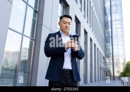 Serious Asian businessman standing on the street near the office with a mobile phone, typing a message, making a call, waiting for an appointment. Stock Photo
