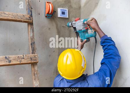 Back photo of the helmeted electrician using the drill to make a hole in the wall. Stock Photo