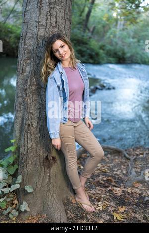 Attractive Young Woman Wearing Denim Jacket Pastel Colors Leaning on Tree Creek Hiking Trail Stock Photo