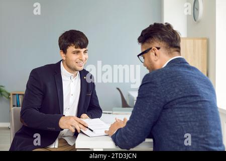 Two business partners in office sign contract sitting at desk, manager gives document to his client. Stock Photo
