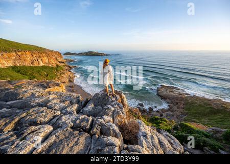 Young woman standing on a rock above the sea in the evening light, beach at The Gap, Robberg Peninsula hiking trail, Garden Route, Robberg Peninsula Stock Photo