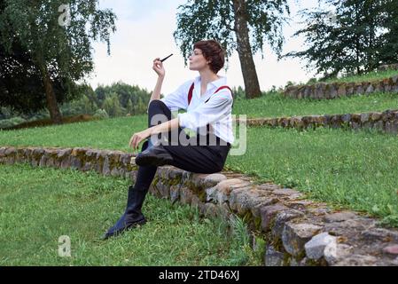 Image of a stylish beautiful woman in a white shirt, black trousers and red suspenders who smokes a pipe. The concept of style and fashion Stock Photo