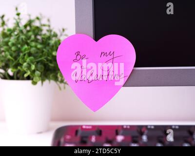 A pink heart shaped paper note with words Be My Valentine on it sticked on to a monitor at an office workplace. Stock Photo