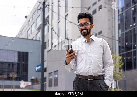 Young successful businessman walking down the street holding a phone in his hands, an Indian man using an application on a smartphone smiling contente Stock Photo