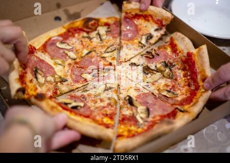 large pizza on the table in a box, cut into pieces, which people take with their hands at the feast. Stock Photo