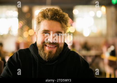Bearded guy programmer or an entrepreneur in glasses in a stylish cafe looks at the camera and smiles. Freelancer works remotely. Online communication Stock Photo
