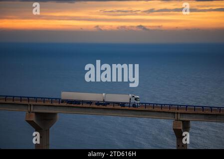Mega-trailer truck driving over a bridge with the sea and dawn sky on the horizon. Stock Photo