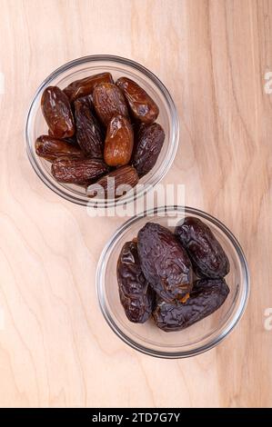 Deglet Nour and Medjool dates, in glass bowls, on a wooden panel, from above. Sun-dried and sweet cultivars of dates. Stock Photo