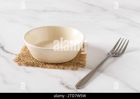 Empty beige bowl and metal fork over marble tabletop. Clean porcelain ramekin for food design mockup. Ceramic crockery and tableware concept. Stock Photo