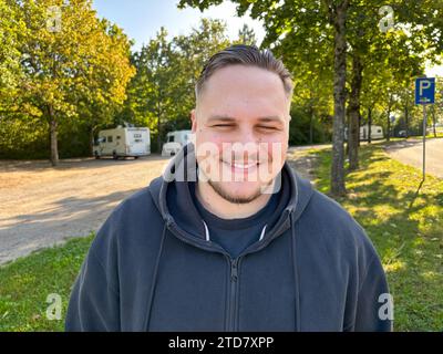 Friendly portrait of a young man smiling with eyes closed wearing a hooded top and black shirt standing in a park near trees Stock Photo