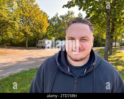 Friendly portrait of a young man wearing a hooded top and black shirt standing in a park near trees Stock Photo