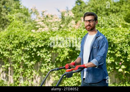 Smiling gardener in shirt mowing lawn with modern lawn mower in summer season. Side view of brunet man in gloves posing, pushing handle of lawn mower, Stock Photo